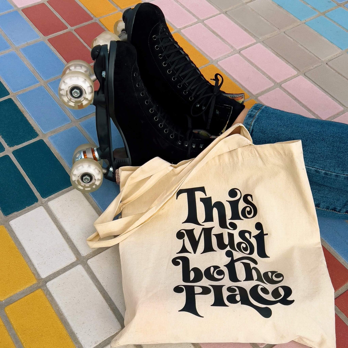 model wearing black roller skates on rainbow tile floor with this must be the place tote bag