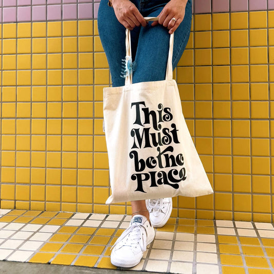 model leaning against yellow tile wall holding this must be the place tote bag